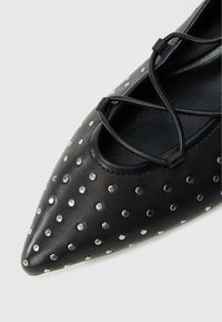 Μπαλαρίνα από μαύρο δέρμα με μυτερή μύτη, διακοσμητικά ασημί καρφιά και λεπτομέρεια με λουριά που διασταυρώνονται. Ομαλή υφή με μιν�ιμαλιστικό σχεδιασμό.