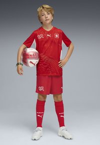 Uniforme de fútbol rojo con acentos blancos, con un patrón texturizado en la camiseta. El balón muestra "NATI", con un diseño en rojo y blanco.