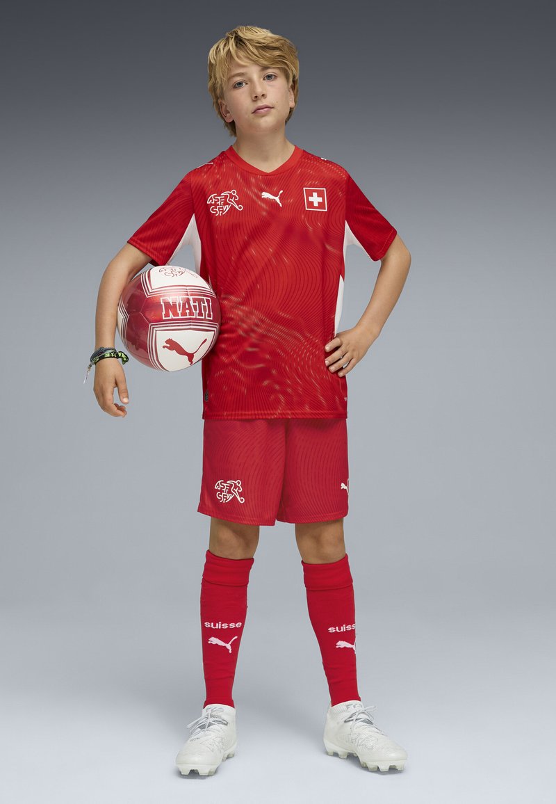 Uniforme de fútbol rojo con acentos blancos, con un patrón texturizado en la camiseta. El balón muestra "NATI", con un diseño en rojo y blanco.