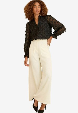 Donna con capelli ricci che indossa una blusa nera a pois trasparente, pantaloni a vita alta e gamba larga crema, tacchi neri e orecchini a cerchio dorati.