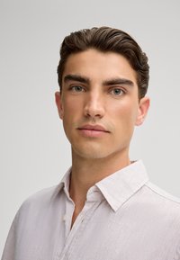 Camicia di lino chiaro e textured con colletto bottone, indossata da un modello maschile con capelli corti e stilizzati e pelle chiara su uno sfondo neutro.