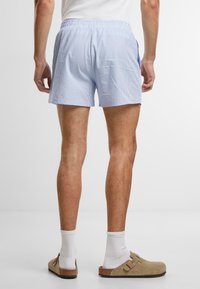 Shorts rayés bleu clair avec une taille élastique et des poches latérales, fabriqués à partir d'un tissu léger. Portés avec des chaussures à enfiler beiges.