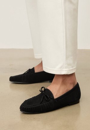 Mocassins loafers en daim noir avec détail nœud portés avec un pantalon blanc longueur cheville sur un sol poli.