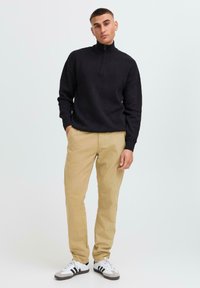 Pull en maille noir avec un col zippé court, associé à un chino beige et des baskets blanches avec des rayures noires. Design simple et minimaliste.