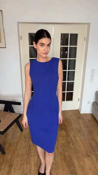 Žena s tmavými vlasy staženými do elegantního drdolu, oblečená v bezrukávých šatech královské modré barvy, stojící na dřevěné podlaze v moderním pokoji se dveřmi se skleněnými panely.