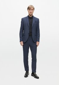 Námorně modrý přizpůsobený oblek se slim fit, strukturovanou tkaninou, na dva knoflíky a odpovídající kalhoty, nošený přes černou košili. Černé mokasíny dotvářejí vzhled.