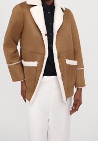 Hnědá svrchní bunda s bílým fleecovým límcem a kapsami, s uzávěrem na knoflíky a kontrastními texturami. Nositá přes černou košili.
