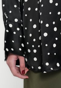 Fekete pöttyös blúz fehér körmintákkal, hosszú ujjal és sima szatén textúrával. Látható alj részlet és egy kéz, amely a szövetet tartja.