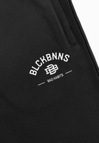 Zwarte sweatpants van zacht materiaal met een witte geprinte logo dat "BLCKBNNS" en "BAD HABITS" in een gebogen ontwerp toont.