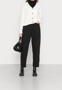 Cárdigan blanco con relieve y botones marrones, combinado con pantalones negros de pierna recta y botas negras de tobillo. La modelo sostiene un pequeño bolso negro.