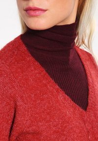 Maglione rosso con texture e scollo a V, indossato sopra un dolcevita bordeaux a coste. I capi presentano diverse trame di lavorazione a maglia e finiture lisce.