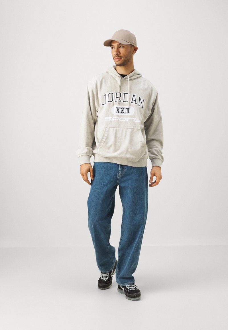 Hellgrauer Hoodie mit "Jordan"-Druck und Logo, kombiniert mit locker sitzenden blauen Jeans und schwarzen Sneakern, ergänzt durch eine beigen Kappe.