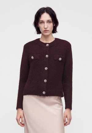 Kvinde med kort mørkt hår bærer en mørkebrun strikket cardigan med dekorative knapper og et lyst beige skørt mod en ensfarvet baggrund.