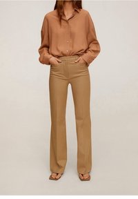 Chemise à manches longues marron clair avec boutons, associée à un pantalon beige taille haute, évasé. Porté avec des sandales plates marron.