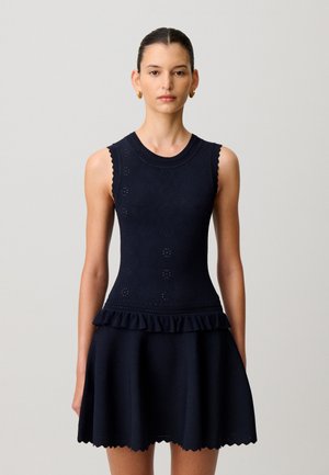 Donna con capelli tirati all'indietro che indossa un vestito blu navy senza maniche con fascia e gonna arruffate, e orecchini a cerchio dorati, in piedi su uno sfondo semplice.