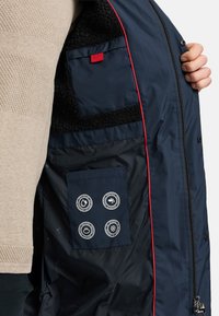 Binnenkant van een marineblauwe jas met een zachte zwarte voering. Voorzien van rode accenten, een zak en vier gelabelde ronde symbolen die water-, wind-, warmte- en sneeuwbestendigheid aangeven.