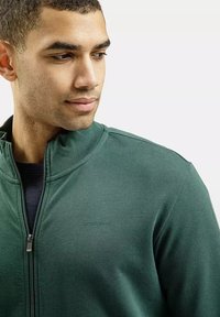 Zielona bluza z zamkiem błyskawicznym, z prążkowanym kołnierzem i logo. Miękka tekstura i swobodny fason, z fasonem blisko ciała i neutralnym tłem.