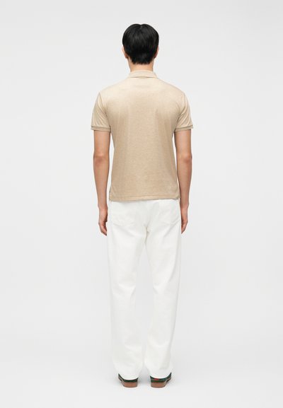 Άνδρας που στέκεται πλάτη, φορώντας μπεζ πόλο μπλουζάκι με κοντά μανίκια, λευκό παντελόνι και καφέ παπούτσια σε απλό λευκό φόντο.