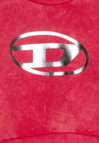 Crveni pamučni dukserica s teksturiranim završetkom i velikim metalik srebrnim ovalnim logotipom u sredini. Okrugli izrez i rebrasti rubovi.