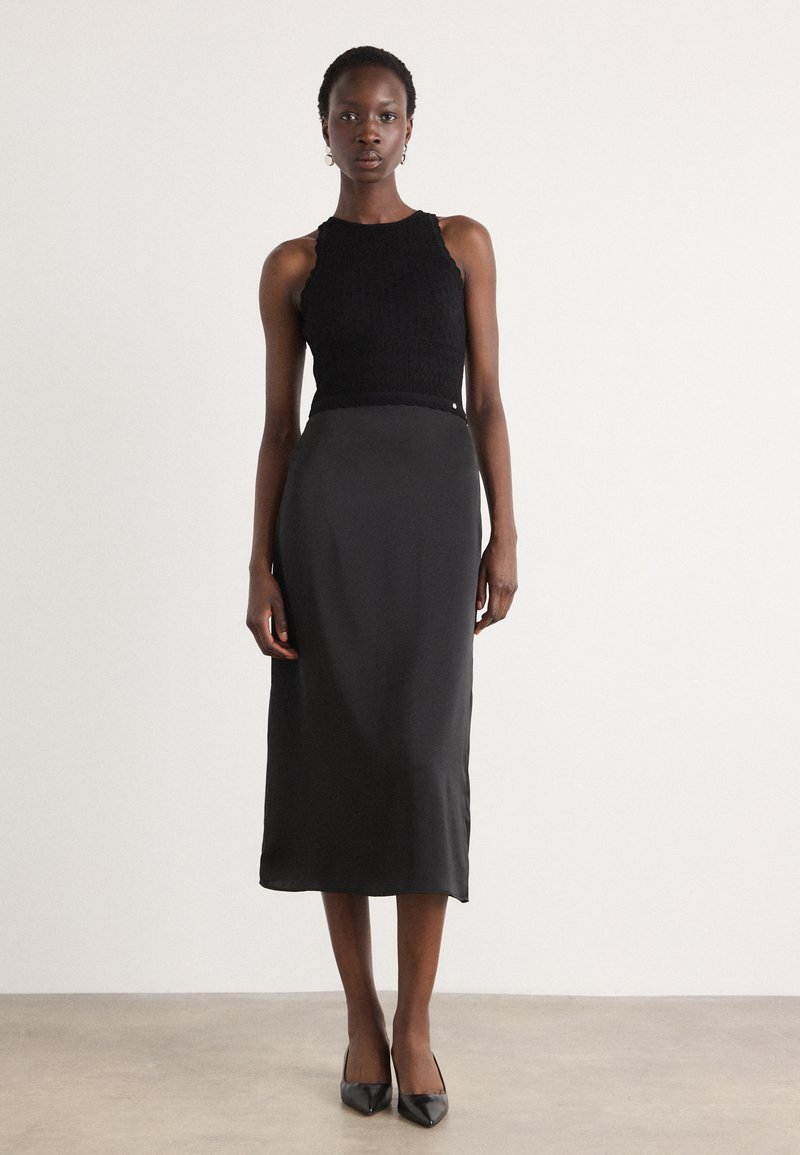 Débardeur en tricot noir à col haut associé à une jupe midi en satin noir lisée. Le modèle porte des talons aiguille noirs.