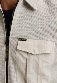 Lichtgrijze zip-up jas met een gestructureerde stof met een geruit patroon. Bevat een voorzak en een merklabel in zwart en geel.