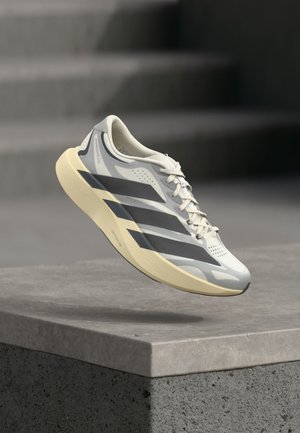 Chaussure de course gris clair et blanc avec des rayures gris foncé et une semelle beige, suspendue au-dessus d'un rebord en béton avec des escaliers flous en arrière-plan.