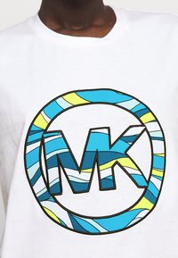 Biała koszulka z okrągłym, niebiesko-żółtym paskowanym logo z literami "MK" na środku. Wzór ma haftowany kontur.