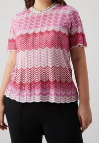 Růžový a bílý pletený top s chevronovým vzorem, s okrajem ve tvaru vlnky a krátkými rukávy, má texturovaný povrch a mírný lesk.