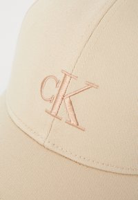Beige baseball cap laget av teksturert stoff med en lett buet skygge, som har en lys rosa brodert "ck" logo på forsiden.