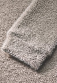 Tessuto grigio chiaro con texture morbida e pelosa, che mostra una leggera lucentezza. Il bordo è leggermente sfilacciato, aumentando la sua qualità tattilità.