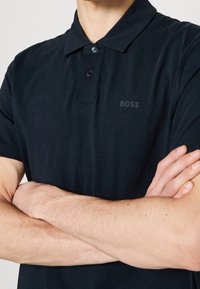 Homme aux bras croisés portant un polo noir à manches courtes avec deux boutons et le logo "BOSS" sur la poitrine.