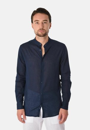 Uomo con capelli scuri corti e barba, indossa una camicia blu scuro con collo alla mandarino abbottonata e pantaloni bianchi su uno sfondo semplice.