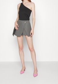 Haut noir côtelé à une épaule, associé à une jupe portefeuille grise avec une poche avant et des accessoires argentés. Le mannequin porte également des chaussures slingback roses.