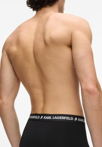 Czarne bokserki z białym logo w pasie, o gładkiej fakturze materiału. Widok z tyłu ukazuje muskularną sylwetkę.