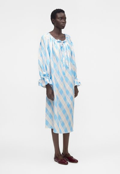 Γυναίκα που φοράει ένα φαρδύ, μέχρι το γόνατο φόρεμα με μπλε και άσπρες διαγώνιες ρίγες και βολάν στα μανίκια, συνδυασμένο με σκούρα κόκκινα παπούτσια τύπου Mary Jane.