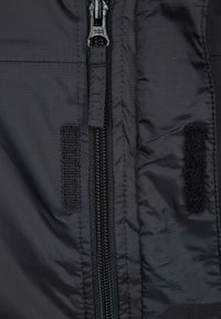 Tela de chaqueta negra que presenta un cierre central con un tirador YKK, junto a dos secciones de velcro para el cierre o ajuste.