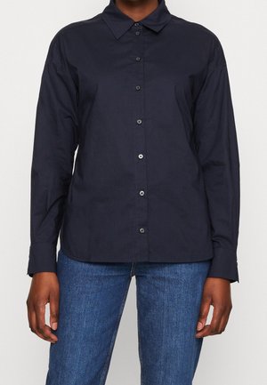 Person iført en mørkeblå skjorte med knapper og lange ærmer, kombineret med blå denimjeans, stående mod en ensartet lys baggrund.