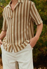Brązowo-kremowa koszula w paski z krótkim rękawem, o fakturowanym dzianinowym wzorze i guzikami, zestawiona z jasnymi spodniami.
