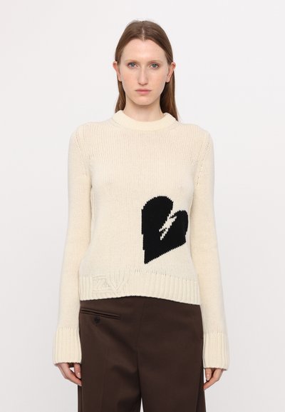 Pull en tricot crème avec un motif de cœur noir à l'avant, col et poignets côtelés, présentant une texture subtile et un logo en bas.
