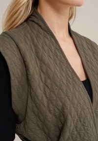 Tikattu oliivinvihreä liivi, jossa on v-pääntie ja teksturoitu timanttikuvio, yhdistettynä tummanväriseen yläosaan. Kangas näyttää pehmeältä ja lämpimältä.