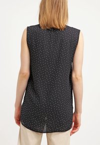Černý bezrukávový top vyrobený z lehkého materiálu, zdobený bílými puntíky a s delším zadním spodním lemem se bočními rozparky.