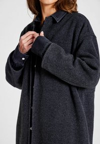 Črna overshirt iz mešanice volne z teksturirano površino, ki ima kontrastni črni usnjeni ovratnik in zapiranje na gumbe, dolge rokave in ohlapen kroj.