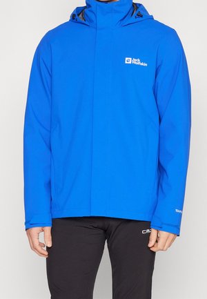 Uomo che indossa una giacca con cappuccio Jack Wolfskin di un azzurro brillante con pantaloni neri, in piedi davanti a uno sfondo chiaro e uniforme.