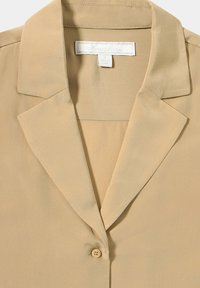Camicia beige a bottoni con un tessuto leggero e liscio, colletto a punta e chiusura a un bottone al colletto.