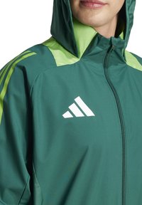 Grüne Sportjacke mit Kapuze, ausgestattet mit hellgrünen Akzenten und drei weißen Streifen auf der Schulter. Reißverschluss vorne, leichtes Material.
