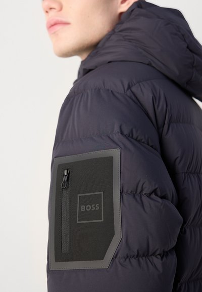Veste matelassée marine avec capuche, dotée d'une poche zippée sur la manche gauche et d'un patch texturé noir. Design léger et isolé.