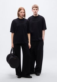 Μαύρα υπερμεγέθη μπλουζάκια με τη φράση «BROKE SINCE MILANO» τυπωμένη στο κέντρο, σε συνδυασμό με μαύρο παντελόνι με ευρύ πόδι και στρογγυλή μαύρη τσάντα.