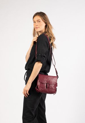 Burgunderfarbene Ledertasche mit verstellbarem Tragegurt, Klappenverschluss und subtiler Textur, gehalten von einem Modell in schwarzer Kleidung.
