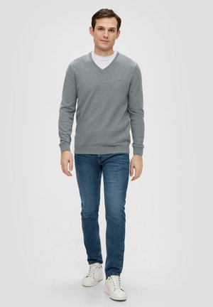 Grijze V-hals trui van zacht materiaal met lange mouwen, gecombineerd met blauwe jeans en witte sneakers, staand tegen een effen achtergrond.