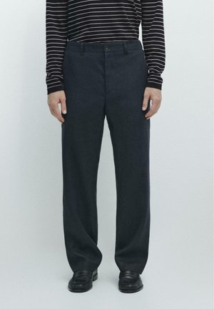 Mężczyzna w czarnych skórzanych butach, luźnych ciemnych spodniach i czarnej koszuli z długim rękawem oraz białymi poziomymi paskami, stojący na białym tle.